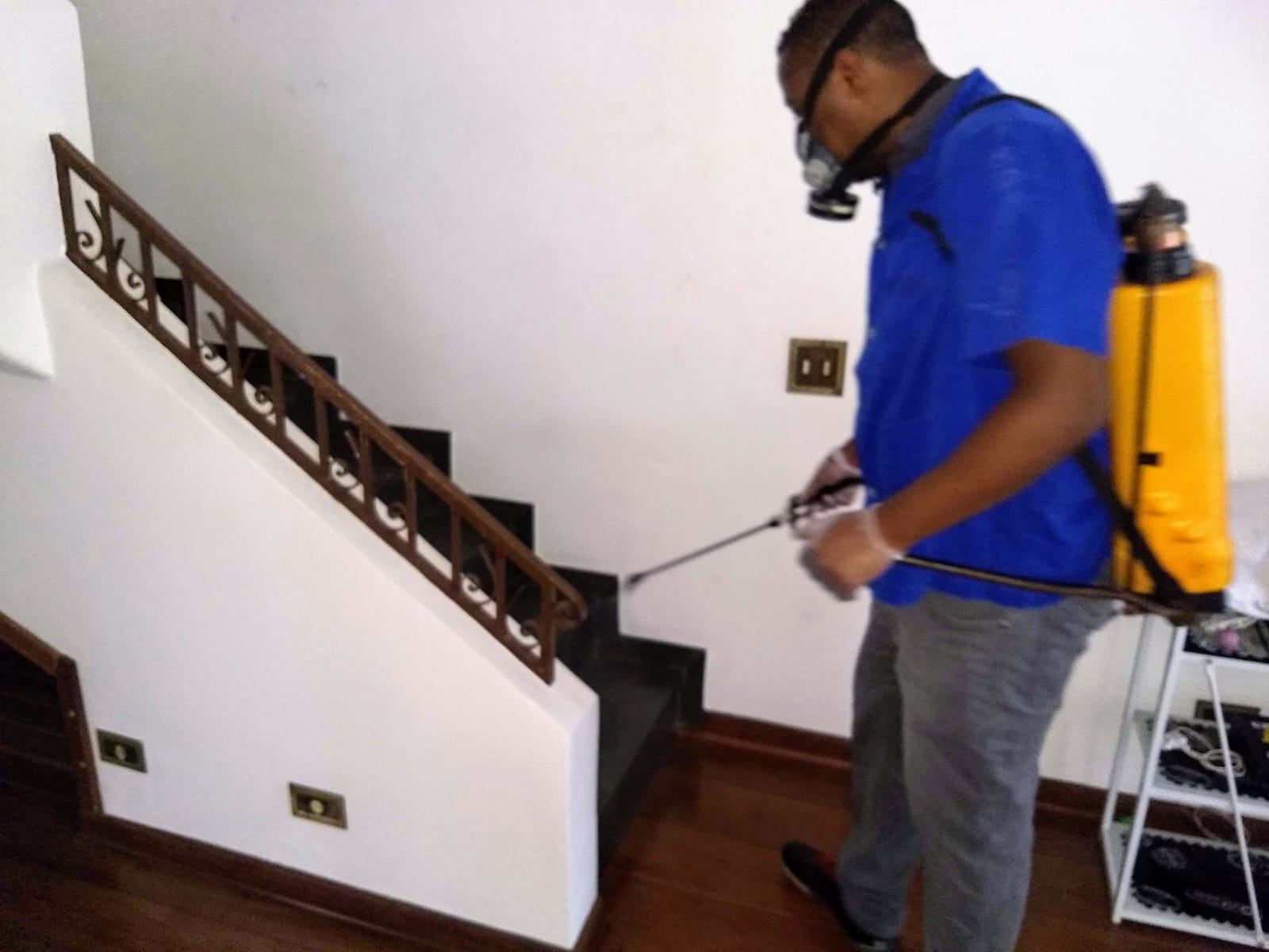Técnico da GMs Controle de Pragas Urbanas, de camisa azul, máscara e pulverizador costal, dedetizando a escada de uma residência em São Paulo